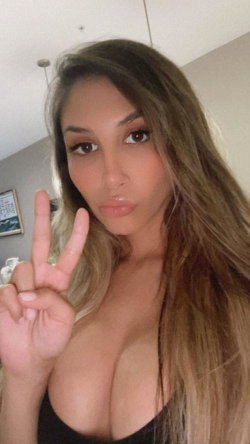 A woman with long brown hair and a curvy build poses in a black top, making a peace sign in a bedroom setting.