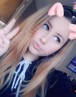 Blonde woman with cat filter taking a selfie, wearing a dark hoodie with white drawstrings, making a peace sign indoors.