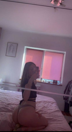 A curvy woman with long hair takes a mirror selfie in a bedroom, wearing a black top and thong while sitting on a bed.