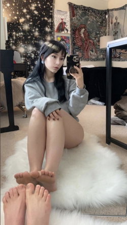 A young woman with long dark hair and bangs sits barefoot on a white rug, taking a mirror selfie in a gray sweatshirt;