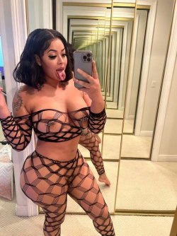 Curvy woman with dark hair in a black mesh bodysuit takes a mirror selfie, sticking out her tongue in a well-lit room