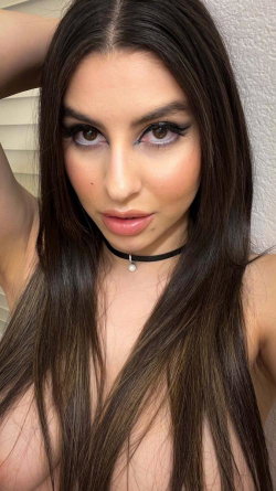Brunette woman with long hair and makeup, wearing a choker, posing topless in a close-up selfie against a textured wall.