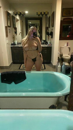 Blonde woman in black lingerie takes mirror selfie in bathroom with turquoise bathtub and dark vanity.