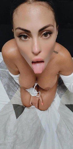 Brunette woman with defined features, wearing white lingerie, posing on a patterned bedsheet with tongue out and arms