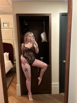 Blonde woman in a leopard-print lace bodysuit takes a mirror selfie in a bedroom, showing tattoos on her legs and arms.