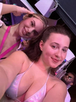 Two women in sparkly tops smile closely together in a dimly lit setting, one taking a selfie with the other leaning in.