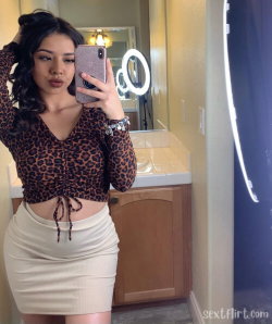 A woman with dark curly hair takes a mirror selfie in a leopard-print crop top and beige mini skirt, standing in a