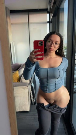 Curvy woman with curly hair and glasses takes mirror selfie in blue ribbed top and low-slung jeans, holding red phone.
