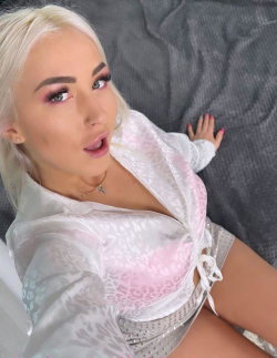 Blonde woman with platinum hair lies on a gray bed, wearing a sheer white lace top and patterned shorts, looking up at