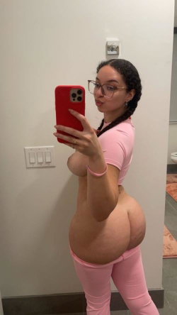 Curvy woman with braided hair and glasses takes a mirror selfie in pink crop top and leggings, showing bare midriff.