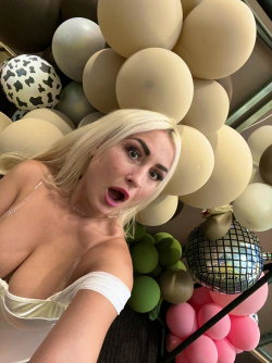 Blonde woman in white lingerie takes a selfie with colorful balloons and a disco ball in the background.