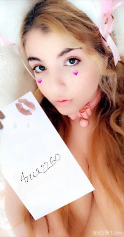 Blonde woman with cat ears and pink choker holds a note with lips and name, bare-chested in a close-up selfie.