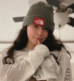 A young woman with long dark hair wears a gray beanie and loose sweatshirt, posing indoors with hand near her chin.