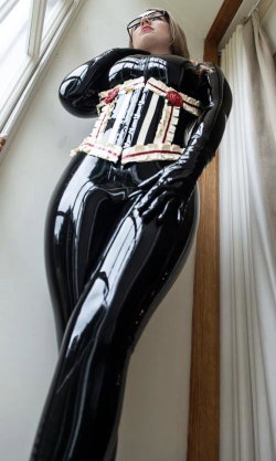 A woman in a shiny black latex bodysuit with a white and red corset detail, wearing gloves and glasses, posing indoors