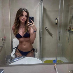 A woman with long brown hair takes a mirror selfie in a bathroom, wearing a dark blue bikini and showing tattoos on her