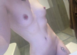 Close-up of a person's torso showing bare breasts, navel piercing, and a small tattoo on the hip, taken in a bathroom