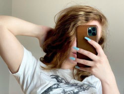 Woman with wavy brown hair taking a mirror selfie in a white graphic tee, hand behind head, holding phone with