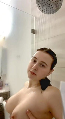 A woman with dark hair takes a selfie in a shower, topless, with water falling from a rain showerhead.