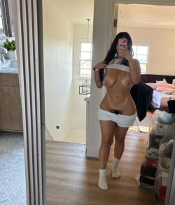 A woman with long dark hair stands in a bedroom taking a selfie in a mirror.
