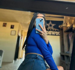 A woman with long dark hair in a blue crop top and jeans takes a mirror selfie, partially obscuring her face with her
