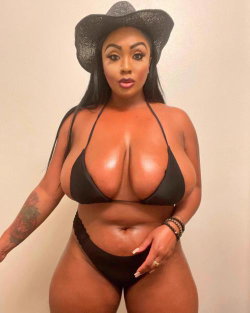Curvy woman with long dark hair wearing a black cowboy hat and matching bikini, posing indoors against a plain wall.