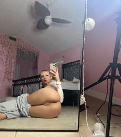 A curvy woman with blonde hair takes a mirror selfie in a pink bedroom, wearing a white long-sleeve top and gray
