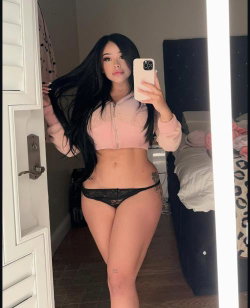 A woman with long black hair takes a mirror selfie in a pink crop hoodie and black lace thong, standing in a bedroom.