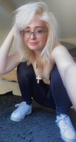 Blonde woman with glasses crouching on a rug, wearing black leggings and white sneakers, topless with a cross necklace