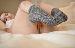 A woman lies on a bed, partially covered by a blanket, wearing gray knitted socks, with her upper body exposed.