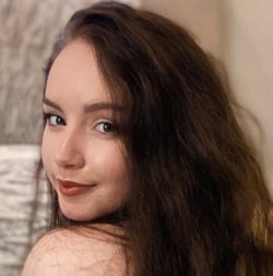 A woman with long dark hair smiles over her shoulder, showing bare shoulders in a softly lit indoor setting.