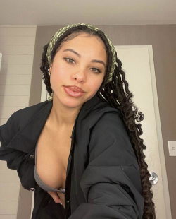 A woman with long braided hair and a patterned headband poses in a black jacket, slightly unbuttoned to reveal a gray