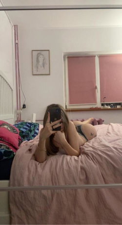 A woman with shoulder-length hair lies on a bed in a bedroom, taking a mirror selfie while wearing a dark bra and pink
