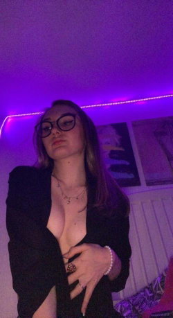 A woman with long dark hair and glasses wears a black blazer open over her chest, posing in a room with purple LED