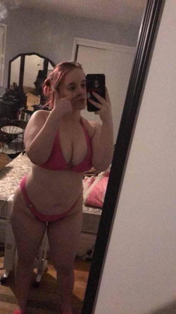 Curvy woman with pink hair in a matching pink bikini takes a mirror selfie in a bedroom, holding her phone with one