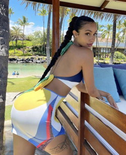 Curvy woman in colorful bikini posing by a wooden cabana, long braided hair, tropical beach background.