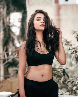 Brunette woman with long dark hair wearing a black crop top and high-waisted bottoms, standing outdoors near foliage.