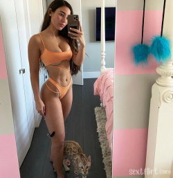A curvy woman in an orange bikini takes a mirror selfie in a pink and white bedroom, with a cat at her feet.