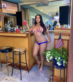 Curvy woman in red-white striped bikini top and blue thong stands barefoot at a tiki bar, holding a drink.