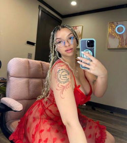 A woman with braided hair and glasses takes a mirror selfie in a red sheer dress, showing tattoos on her arm and