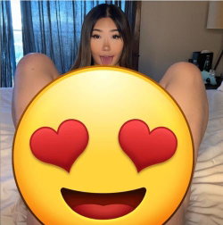 Asian woman with long dark hair lying on a bed, legs spread, sticking tongue out, holding a heart-eyes emoji over her