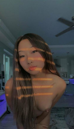 Asian woman with long dark hair, topless, posing indoors with orange light stripes across her body in a dimly lit room.