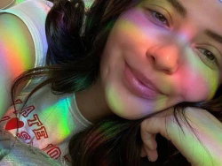 A smiling woman with long dark hair rests her head on her hand, wearing a white tank top under colorful rainbow