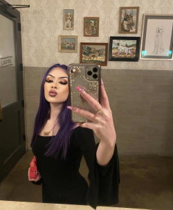 A woman with purple hair and dramatic makeup takes a mirror selfie in a black top, holding a decorated phone in a room