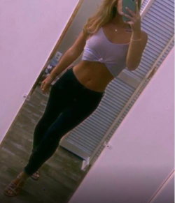 Blonde woman in a white crop top and black leggings takes a mirror selfie, showing toned midriff and slim build.