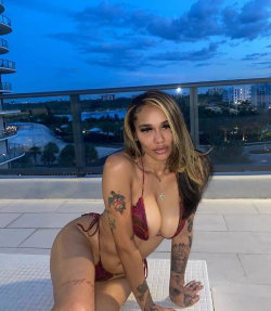 Curvy woman with long ombre hair and multiple tattoos poses in a red patterned bikini on a rooftop terrace at dusk.