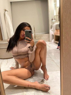 Brunette woman in a beige bodysuit sits on marble floor, taking mirror selfie in bathroom with visible tattoos on legs