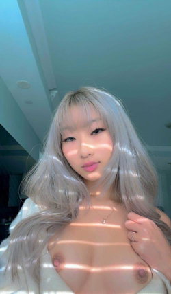 Asian woman with long silver hair and bangs, topless in a white robe, holding her chest in a sunlit indoor setting.