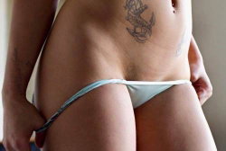Curvy woman in light blue thong with anchor tattoo on lower abdomen, hands adjusting underwear.