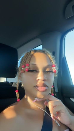 A woman with braided hair adorned with pink beads poses in a car, wearing a dark top and delicate necklace, hand near