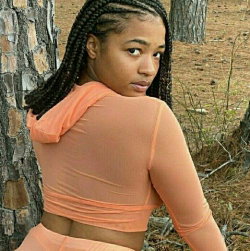 A woman with braided hair in a peach-colored sheer crop top and matching bottoms, standing outdoors near trees.